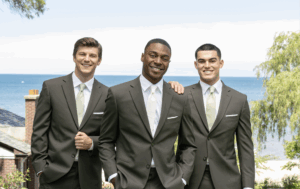 Three men in matching dark suits with light green ties stand outdoors, smiling, with a scenic view of trees, rooftops, and a large body of water in the background. One man has his hand on another’s shoulder. Should I rent or buy my suit or tuxedo?